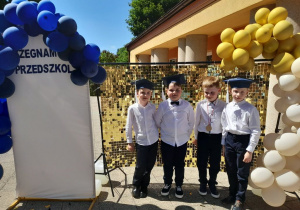 Czterech chłopców pozuje do zdjęcia na tle złotej ścianki ozdobionej balonami.