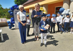 Hubert stoi z dyplomem i pluszakiem na tarasie, a za nim Pani dyrektor, Pani Monika i grupka dzieci.
