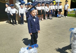 Bartek stoi na środku tarasu i zaprasza na występ. W tle ubrani na galowo rówieśnicy i fragment dekoracji.