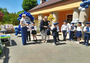 Pani Monika wręcza dyplom Wiktorowi, a obok stoi Pani dyrektor i grupka rówieśników.