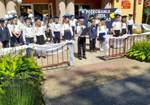 Mikołaj czyta list pożegnalny. Za chłopcem stoją rówieśnicy. W tle budynek przedszkola, dekoracja.