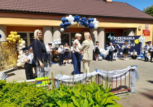 Pani dyrektor wręcza mamie Tosi podziękowanie, a obok stoi Pani Monika. W tle stoją dzieci z dyplomami i upominkami.