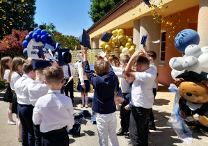Dzieci stoją w małym kole i podrzucają do góry birety.