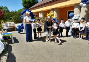 Pani Monika wręcza dyplom Weronice. Obok stoi Pani dyrektor i grupka rówieśników.