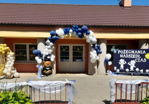 Dekoracja na tarasie przedszkolnym z okazji zakończenia roku szkolnego i pożegnania przedszkola. Nad drzwiami wisi biało-niebieska girlanda z balonów, z prawej strony na granatowym materiale przypięte są dwie postacie absolwentów przedszkola, słoneczko, motyl, białe kwiaty, napis: "Pożegnania nadszedł czas". Z lewej strony znajduje się stojak z balonami, balon - absolwent przedszkola, złota błyszcząca ścianka z dołączonymi po boku balonami w kolorze złotym, kremowym.