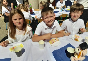 Troje dzieci siedzi przy stole i degustuje tort.