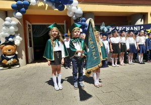 Amanda, Mateusz i Zuzia stoją z proporcem przedszkola na środku tarasu. W tle dzieci śpiewają hymn przedszkola, dekoracja.
