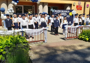 Dzieci w galowych strojach stoją w parach na tarasie. W tle budynek przedszkola oraz dekoracja z okazji pożegnania przedszkola.