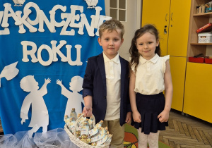 5. Aleksander i Jagoda z uśmiechem na twarzy stoją przy dekoracji.Olek trzyma w ręku koszyk ze słodkimi upominkami dla pracowników przedszkola.