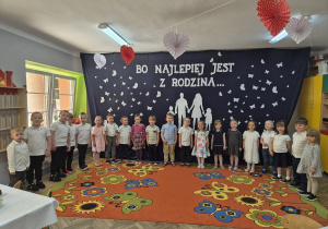 1. Dzieci z grupy "Motylki" stoją w odświętnych strojach na tle dekoracji. Na granatowym napisie widać napis: "Bo najlepiej jest z rodziną…", białe postacie rodziców z dziećmi trzymającymi się za ręce i unoszące się wokół nich białe motyle.