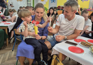5. Józio na kolanach u mamy wręcza tacie upominek w postaci drewnianej ramki z serduszkiem w środku.