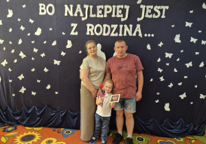 7. Mama, tata i Kacperek stoją na tle dekoracji i pozują do zdjęcia. Kacperek trzyma w dłoniach upominki dl rodziców.