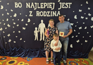 9. Krzyś z rodzicami stoją na tle dekoracji: "Bo najlepiej jest z rodziną…". Mama trzyma w dłoni upominek: rożek z kwiatkami w postaci serc, a tata drewnianą ramkę z czerwonym serduszkiem w środku.