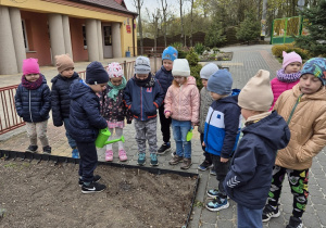 6. Leon stoi trzymając zieloną konewkę i podlewa wcześniej zasiane nasiona kwiatów. Pozostałe dzieci z grupy stoją i czekają na swoją kolej w podlewaniu.