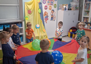 Dzieci poruszają się w kole przy muzyce trzymając chustę animacyjną, na której leżą balony w kropki.