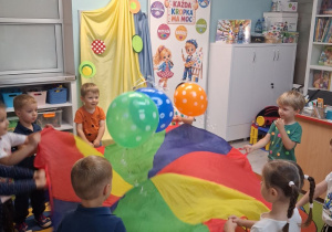 Dzieci stoją przy chuście animacyjnej i podrzucają do góry balony.