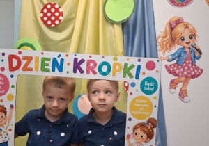 Dwóch chłopców pozuje do zdjęciana tle dekoracji w ramce na której znajdują się kolorowe kropki, dzieci oraz napis „Dzień kropki”.