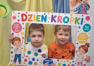 Dwóch chłopców pozuje do zdjęcia na tle dekoracji w ramce na której znajdują się kolorowe kropki, dzieci oraz napis „Dzień kropki”.