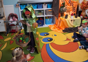 Przedszkolaki siedzą na dywanie i klaszczą w dłonie, a przed nimi stoi dziewczynka przebrana za jesień trzymająca wiklinowy kosz z darami. W tle dekoracja z okazji powitania jesieni.