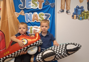 8. Dwóch chłopców pozuje do zdjęcia z balonami w kształcie samochodów na tle dekoracji.