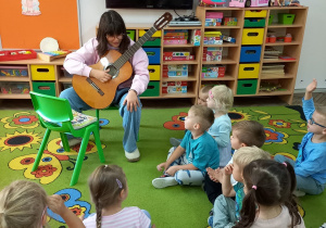 1. Amelka siedzi na krześle trzymając w ręku gitarę. Dzieci słuchają ciekawostek na temat tego instrumentu.