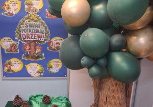 1.Dekoracja z okazji Dnia Drzewa: na tablicy przypięte są ilustracje nt "Świat potrzebuje drzew”, pod tablicą na szafce okrytej zielonym materiałem leżą szyszki. Obok stoi drzewo wykonane z balonów.