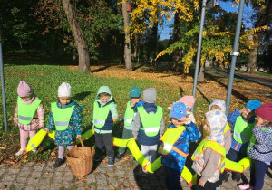 1. Dzieci w kamizelkach odblaskowych trzymają węża i kosz wiklinowy na kasztany. W tle krajobraz jesiennego parku.