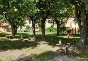 Pomiędzy drzewami stoją urządzenia terenowe: huśtawki, karuzela, lokomotywa, zjeżdżalnia.