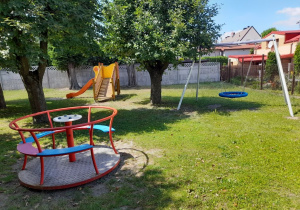 Pomiędzy drzewami stoją: karuzela, zjeżdżalnia i huśtawka "bocianie gniazdo".