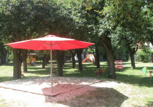 Na pierwszym planie piaskownica z czerwonym, rozłożonym parasolem. W tle pomiędzy drzewami urządzenia terenowe.