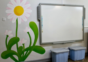 Sala grupy IV "Słoneczka" - na ścianie wisi tablica multimedialna, a obok namalowana jest stokrotka. Pod tablicą stoją dwa pojemniki.