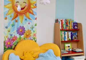 Sala grupy IV "Słoneczka" - na podłodze stoi kanapa, a obok kącik książki. Na ścianie znajduje się dekoracja przedstawiająca słonko, kwiatki, wiszące chmurki oraz napis "Słoneczka".