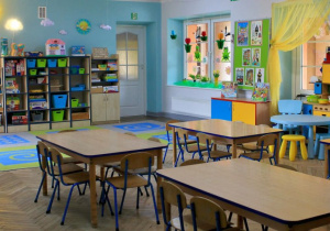 Sala grupy IV "Słoneczka" - na podłodze leży dywan, obok stoją stoliki dla dzieci. Z prawej strony znajduje się kącik domowy, tablica tematyczna, udekorowane okno. Dalej, przy ścianie stoją szafki z układankami.