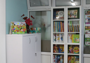 Sala grupy I "Pszczółki" - na szafce w białym kolorze stoi wazon z kwiatami, książka i pudełko z planszami, a obok znajduje się biblioteczka z książkami.