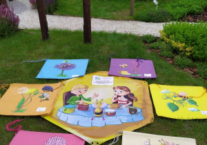 Na trawniku leżą elementy gry edukacyjnej "Gra o miodek": sześć pelerynek w różnych kolorach z symbolami kwiatów roślin miododajnych oraz laminowany baner . W tle elementy wyposażenia części tematycznej "Z wizytą u pszczół".